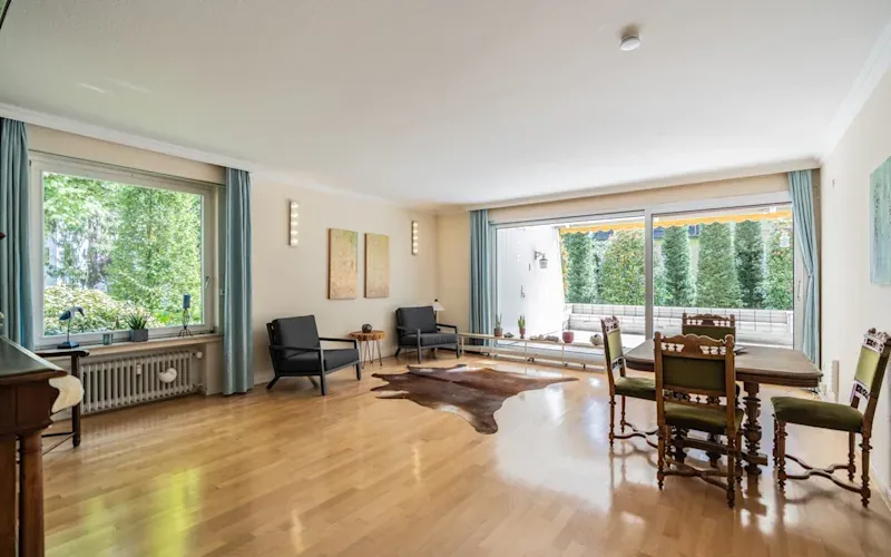 Helles Wohnzimmer mit großen Fenstern, Holzboden, Sitzecke und Esstisch mit Stühlen.