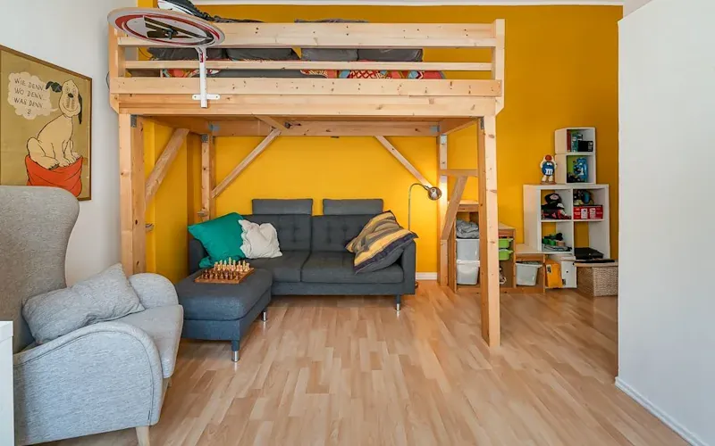 Wohnzimmer mit Hochbett, grauem Sofa, gelber Wand und Regal mit Aufbewahrungsboxen.