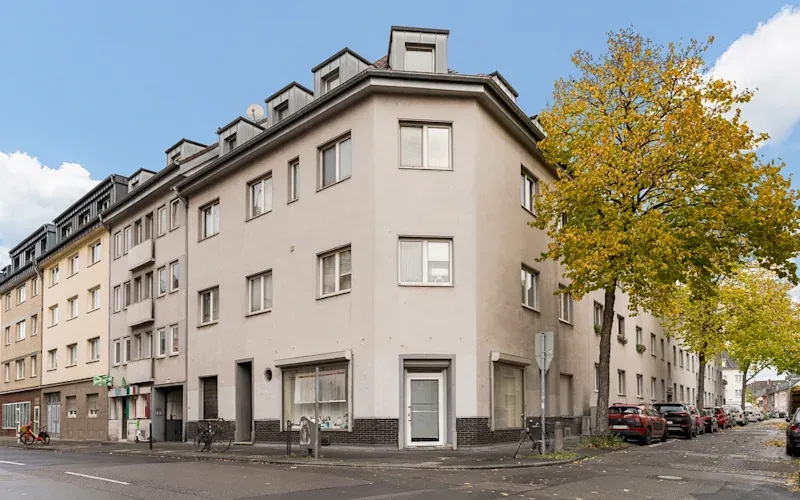 Eckhaus mit mehreren Stockwerken, Fenstern und eingeschossigem Dach, an einer Straßenecke mit Bäumen.