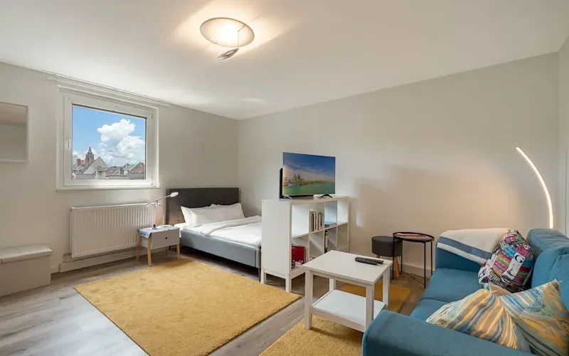 Helles Wohn- und Schlafzimmer mit Bett, Sofa, Fernseher und gelbem Teppich.