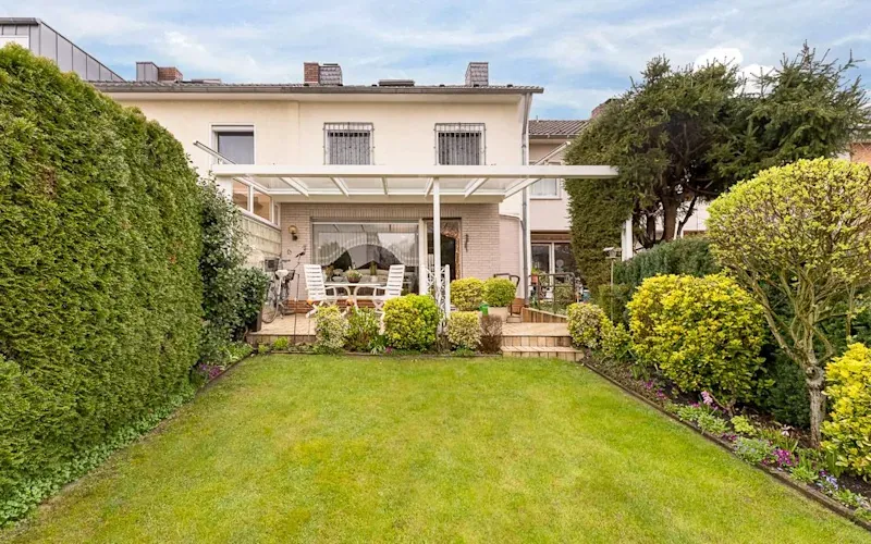 Reihenhaus mit überdachter Terrasse, Garten und Gartenmöbeln, umgeben von Hecken und Büschen.