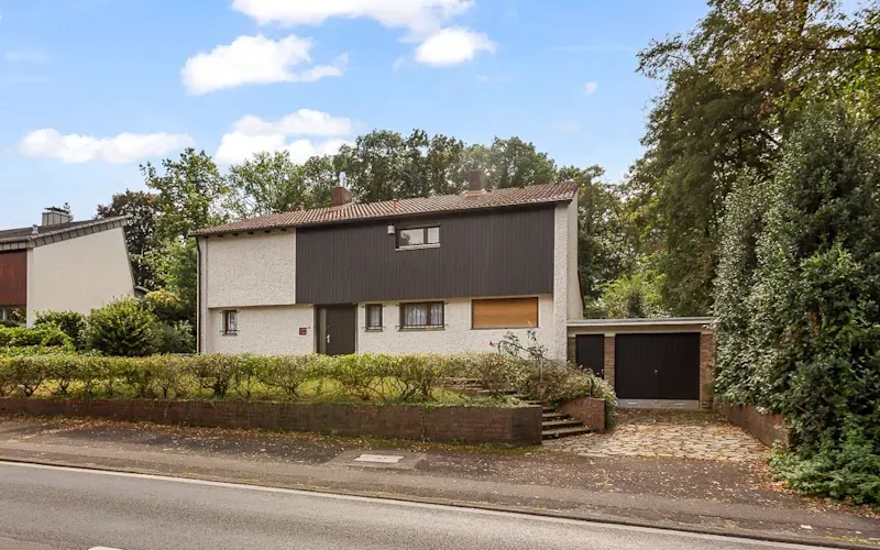Zweistöckiges Wohnhaus mit Garage, Vorgarten und Bäumen im Hintergrund an einer Straße.