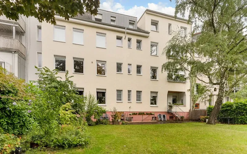 Mehrstöckiges Wohnhaus mit vielen Fenstern, Garten und Bäumen im Vordergrund.