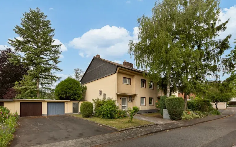Zweistöckiges Wohnhaus mit Vorgarten, großen Bäumen und Garage an einer ruhigen Straße.