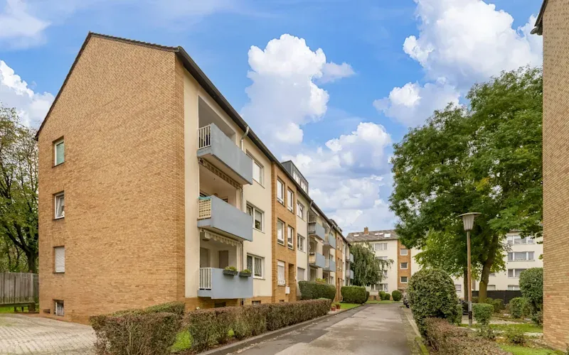 Mehrstöckiges Wohnhaus mit Balkonen, umgeben von Büschen und Bäumen unter blauem Himmel.