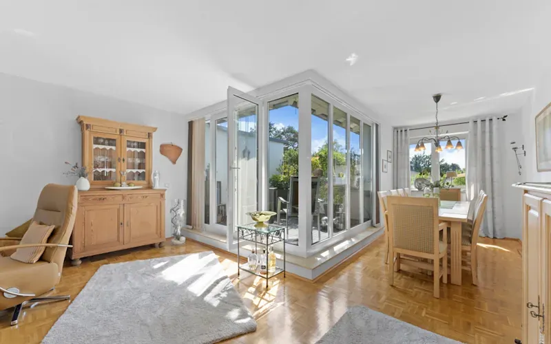 Helles Wohnzimmer mit Essbereich, Holzmöbeln, Teppich und Zugang zu einem Balkon durch große Glasfenster.