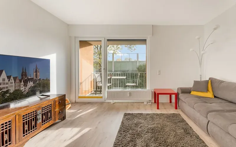 Helles Wohnzimmer mit Sofa, rotem Tisch, Fernseher und Zugang zu einem Balkon mit Stühlen.