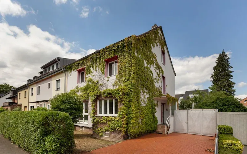Eckhaus mit Efeu bewachsener Fassade, rotem gepflastertem Hof und Hecke am Gehweg.